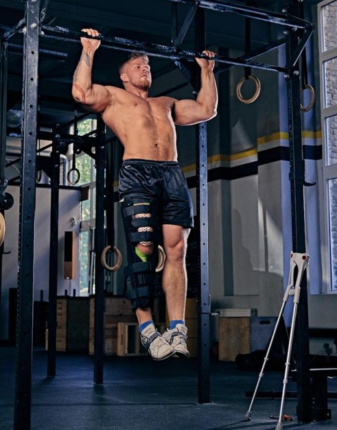 blond bodybuilder with injured leg in a bandage doing pull ups on cross fit stand in a gym.